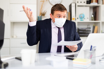 Irritated male in disposable face mask working in business office using laptop