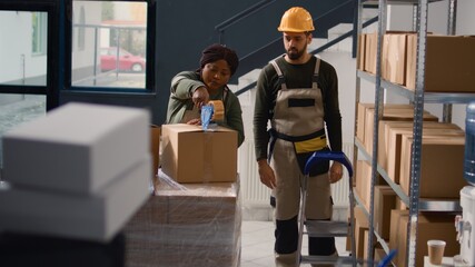 Manager wearing protective equipment in warehouse supervising african american trainee sealing cardboard box packages. Storage area personnel properly securing goods to avoid problems during shipping - obrazy, fototapety, plakaty