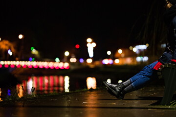 Wyprostowane nogi osoby siedzącej na ławce na tle świateł miasta nad rzeką, kolorowy bokeh...