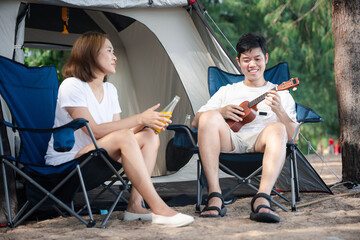 Camping bliss, A cheerful Asian couple, by their tent, serenades each other with ukulele tunes,...