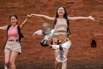 two asian tourist girl happy run jump and pointing hand while the pigons fly and eat at the tha...