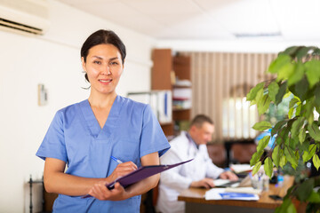Portrait of polite Asian female doctor wearing blue uniform meeting patient in clinic, filling out...
