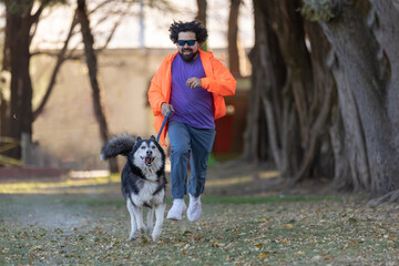 Naklejka na ściany i meble Young Latino man with sunglasses, hair and beard running with his husky dog in park