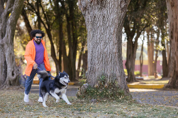 Naklejka na ściany i meble Young Mexican man with curly hair walking his husky dog in park