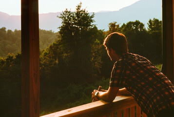 A young man in a flannel shirt leans on the railing of the porch of a rustic cabin overlooking forests and mountains at sunset near Oak Ridge, Tennessee. He holds a glass of beer in his hands. Closeup