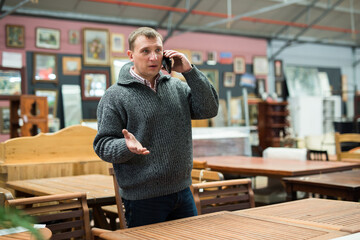 Portrait of young adult man talking by phone while looking for furniture at showroom