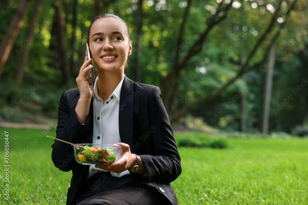 Wall mural Lunch time. Happy businesswoman with container of salad talking on smartphone on green grass in park, space for text