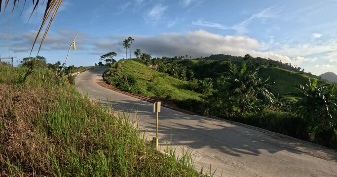 Concrete hillside road curves through green tropical slopes near Bato Ni Ningning in San Vicente Palawan with palms grass and blue sky above the valley