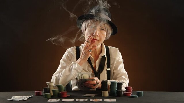An elderly gangster woman smokes a cigarette and holds a stack of money she won in poker. She is dressed in a white shirt with suspenders and a black hat. Cards and chips are on the table. 8K RAW.