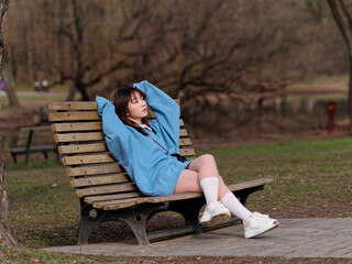 Fototapeta premium Beautiful young Chinese woman posing in sunny summer forest, wearing blue loose oversized top and miniskirt with small shoulder bag. Emotions, people, beauty, youth and lifestyle portrait.