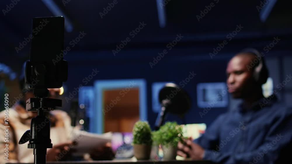 Poster focus on smartphone on tripod recording vlogger show host in blurry background interviewing guest in