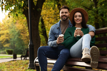 Naklejka premium Happy young couple with cups of coffee spending time together on wooden bench in autumn park, space for text