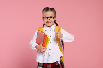 Happy schoolgirl in glasses with backpack and books showing thumb up gesture on pink background