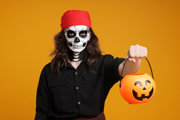 Man in scary pirate costume with skull makeup and pumpkin bucket on orange background. Halloween...