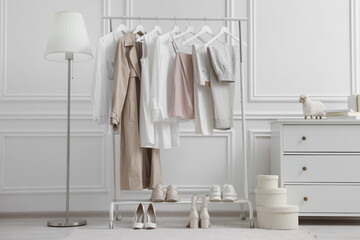 Rack with different stylish women`s clothes, shoes, lamp and dresser near white wall in room