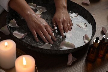 Woman soaking her hands in bowl of water and flower petals at table, closeup. Spa treatment