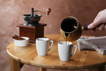 Turkish coffee. Woman pouring brewed beverage from cezve into cup at wooden table, closeup