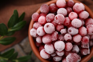 Frozen red cranberries in bowl and green leaves on table, top view