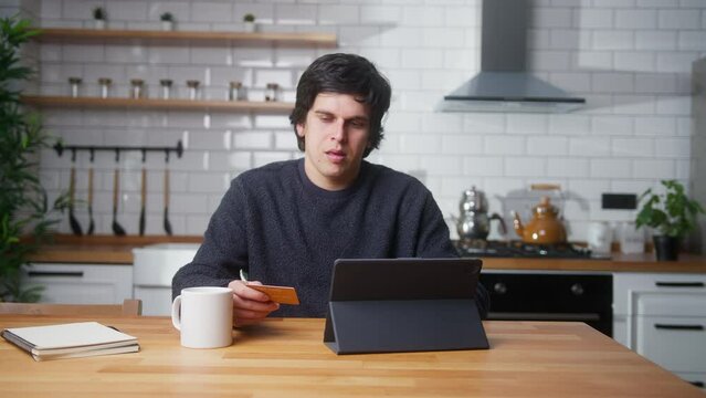 Young man sitting in kitchen at home entering credit card number on tablet device for makes distant goods purchase. Credit card is declined or out of limit, feeling sad and unhappy