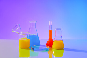 Lab beakers, Erlenmeyer flasks, and spherical glassware hold red, blue, and yellow liquids. A circular glass podium stands on a blue-purple gradient background.