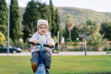 Little smiling girl sits on a swing-balancer, holding the handle, in the park