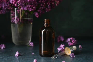 Fototapeta na wymiar Bottle of cosmetic oil with beautiful lilac flowers on dark table