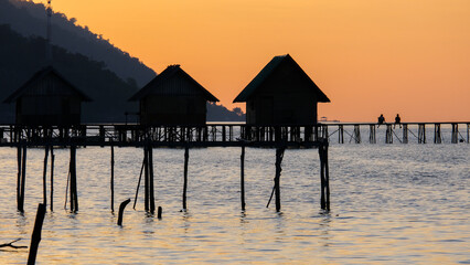 Silhouetted people relaxin on a wharf jetty over ocean water with traditional beach huts against golden orange sunset sky in Raja Ampat, West Papua, Indonesia