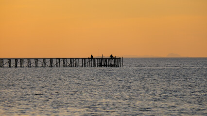 Fototapeta premium Silhouetted people sitting and relaxing on a long wharf jetty over ocean water against a beautiful golden orange sunset sky in Raja Ampat, West Papua, Indonesia