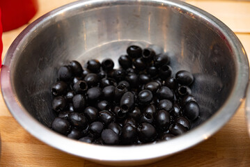 Black Olives in a Metal Bowl on Wooden Surface. Pitted black olives glistening with moisture, contained in a reflective metal bowl set on a wooden kitchen counter.