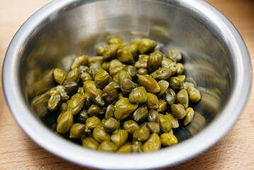 Marinated Capers in Stainless Steel Bowl. Close-up view of pickled green capers in a metallic bowl, a tangy garnish for Mediterranean dishes.