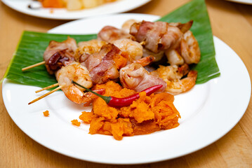 Bacon-Wrapped Shrimp with Mashed Sweet Potato. Bacon-wrapped shrimp skewers over a bed of banana leaf, served with a side of mashed sweet potato and a garnish of red chili pepper.