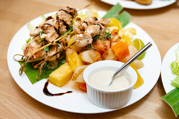 Chicken Skewers with Tropical Fruit Salad and Dip. Grilled chicken skewers served with a colorful tropical fruit salad, accompanied by a creamy dip, presented on a banana leaf-lined plate.