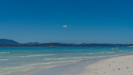 A serene seascape. The waves of the aquamarine ocean roll towards the sandy beach. Boats, yacht in the distance. A mountain range against a clear blue sky. Copy space. Madagascar. Nosy Tanikeli   