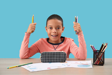 Little African-American boy with pencil case at table on blue background