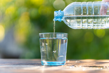Clear, clean water in a glass. Hand holding drinking water bottle pouring into glass on wooden table on blurred green nature background