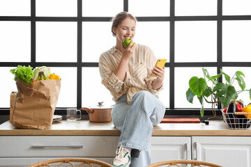 Young woman with apple using mobile phone in kitchen - obrazy, fototapety, plakaty