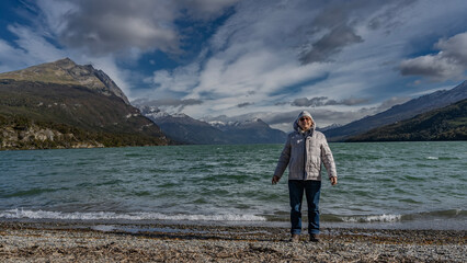 A man in a down jacket and a hat stands on the shore of a beautiful emerald lake, smiling. The waves are foaming on the pebble. Behind is the picturesque snow-capped mountains-Andes. Clouds, blue sky