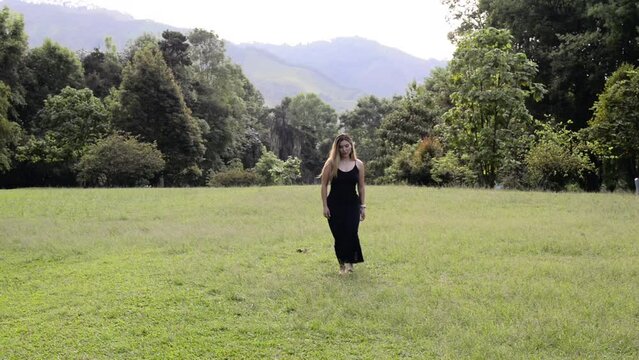 Blonde girl walks on a green grass and black dress without shoes. Wide shot
