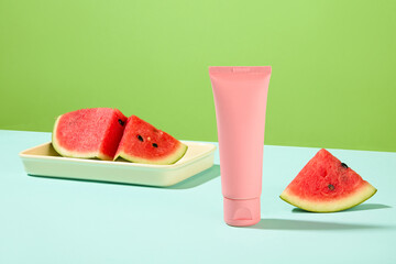 Front view of an unlabeled pink cosmetic tube displayed with watermelon on a blue surface with a green background. Mockup of natural cosmetics.
