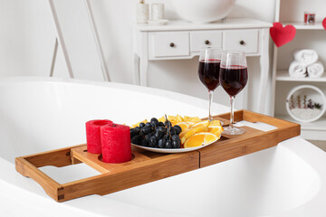 Bathtub tray with candles, fruits and glasses of wine in bathroom. Valentine's Day celebration