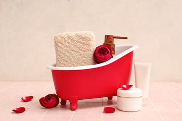 Mini bathtub with cosmetic products, sponge and roses on pink tile near white wall. Valentine's Day celebration