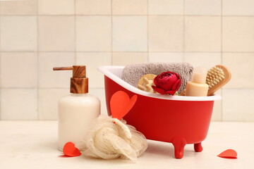 Mini bathtub with bath supplies, paper hearts and rose on white table near tile. Valentine's Day celebration