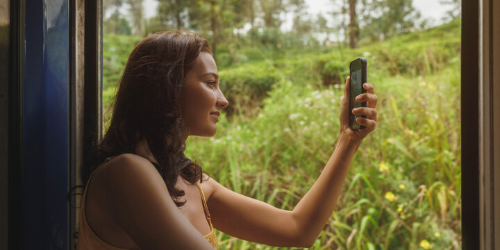 Enjoying travel. Train travel of tourist woman with mobile phone. An attractive brunette woman standing near the wagon open doors using a smartphone, taking a photo of tea plantations, a famous