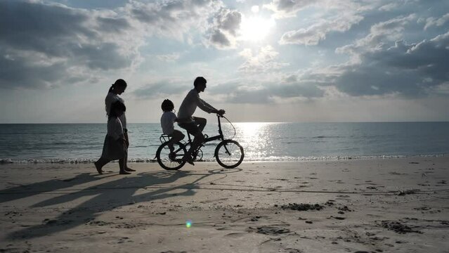 Asian family, father, mother, daughter, and son enjoy riding bicycles and walking along the beach during the sunset, Summer Vacation. Slow Motion