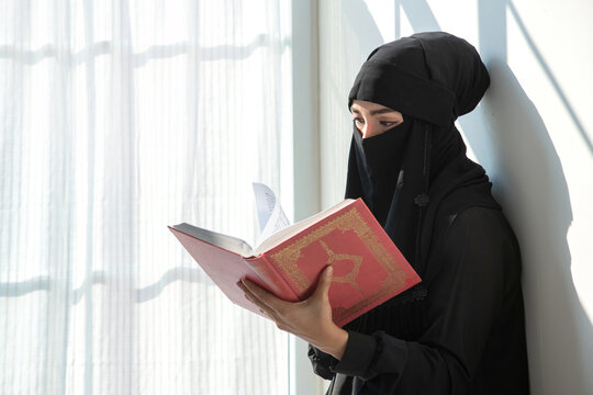 Beautiful Muslim woman wearing black hijab or Nigab studying holy Quran near window at home
