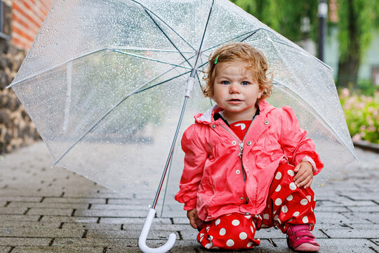 Cute adorable baby girl discovering snail on a walk. Beautiful curly toddler child having fun on rainy day. With big umbrella, child in waterproof clothes