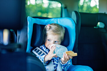 Adorable baby girl with blue eyes sitting in car safety seat. Toddler child going on family vacations and jorney. Smiling happy child during traffic jam, drinking milk from bottle and eating bisquit