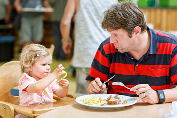 Toddler girl eating healthy vegetables and unhealthy french fries potatoes. Cute happy baby child taking food from parents dish in restaurant. Father eating in fast food restaurant with daughter
