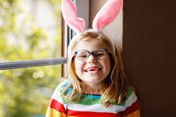 Portrait of a happy little girl with bunny ears looking outside sitting by a window. Easter holiday. Adorable child happy about holiday. - obrazy, fototapety, plakaty
