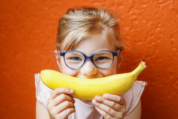 Happy little child girl with yellow banana like smile on orange background. Preschool girl with glasses smiling. Healthy fruits for children © Irina Schmidt